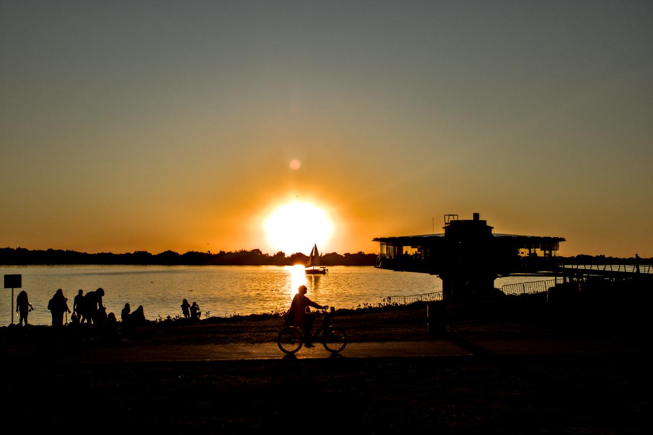 Sol se põe entre bicicleta e veleiro diante de lago.