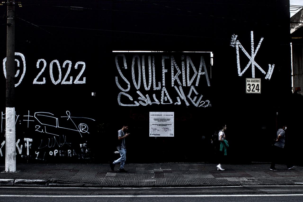 Construção preta pichada em contraste com poste branco e cartaz no portão.