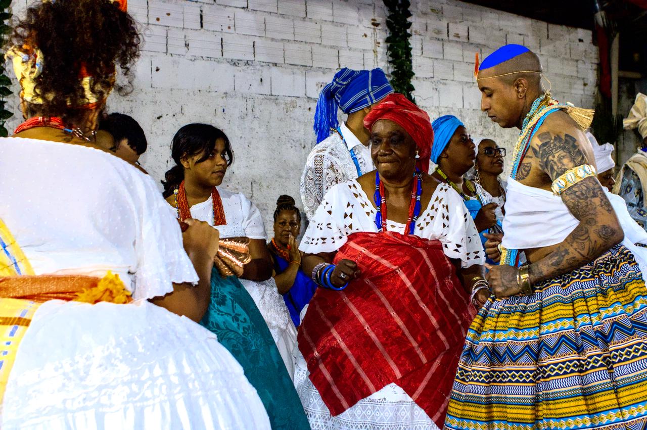 Idosa vestida em vermelho ao lado de jovem Logunedé.