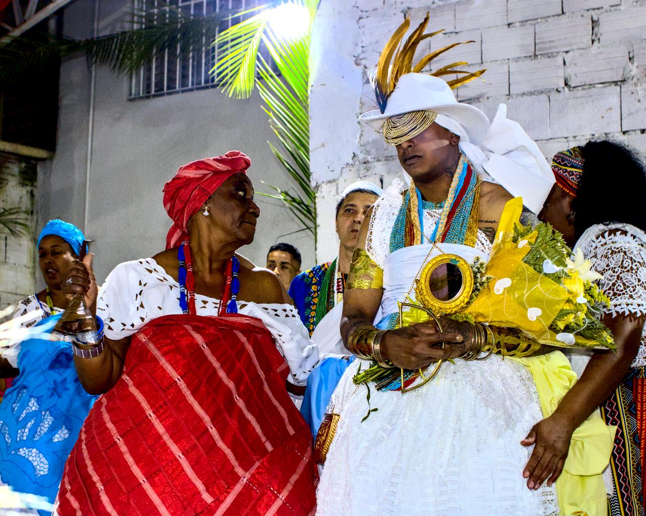 Idosa vestida em vermelho ao lado de jovem Logunedé.