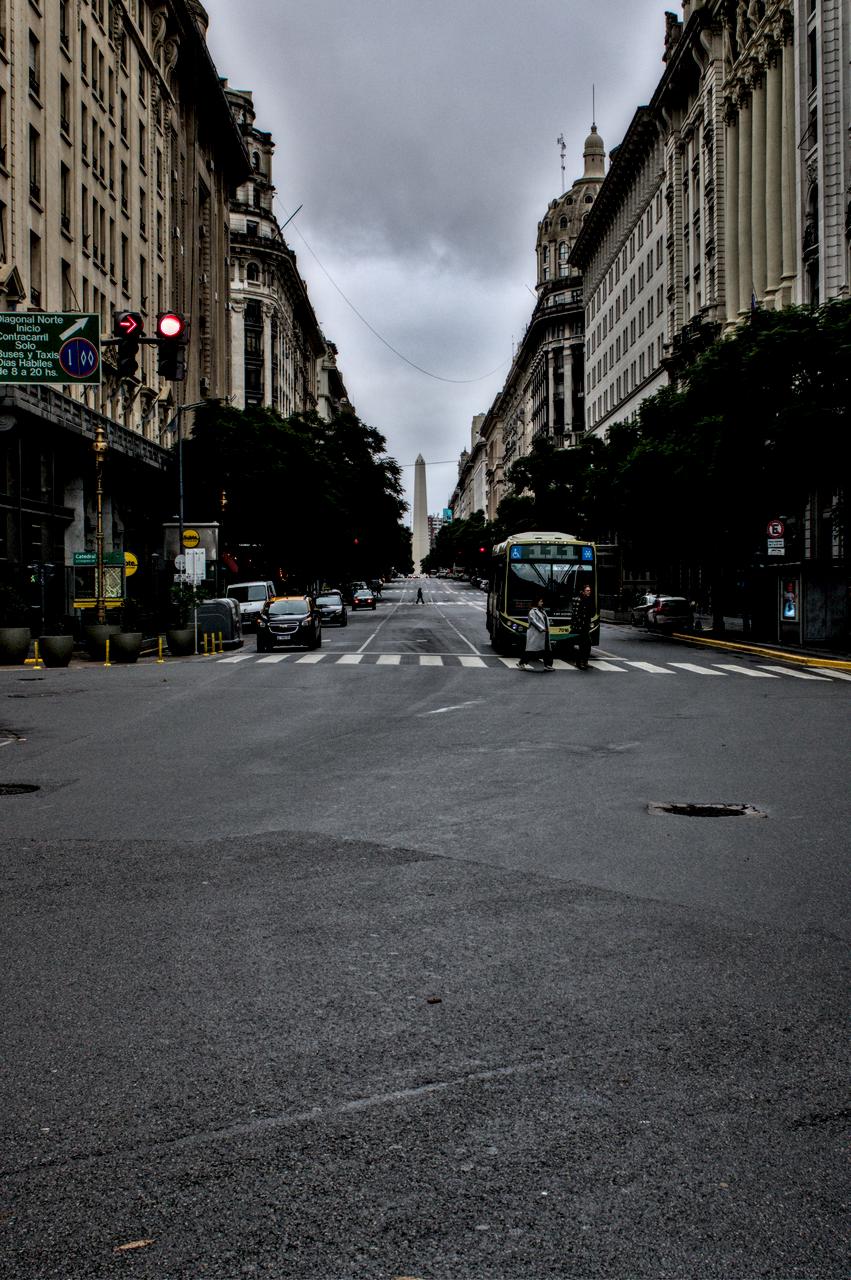 Rua repleta de edifícios e obelisco ao fundo; no meio, pedestre atravessa a rua.