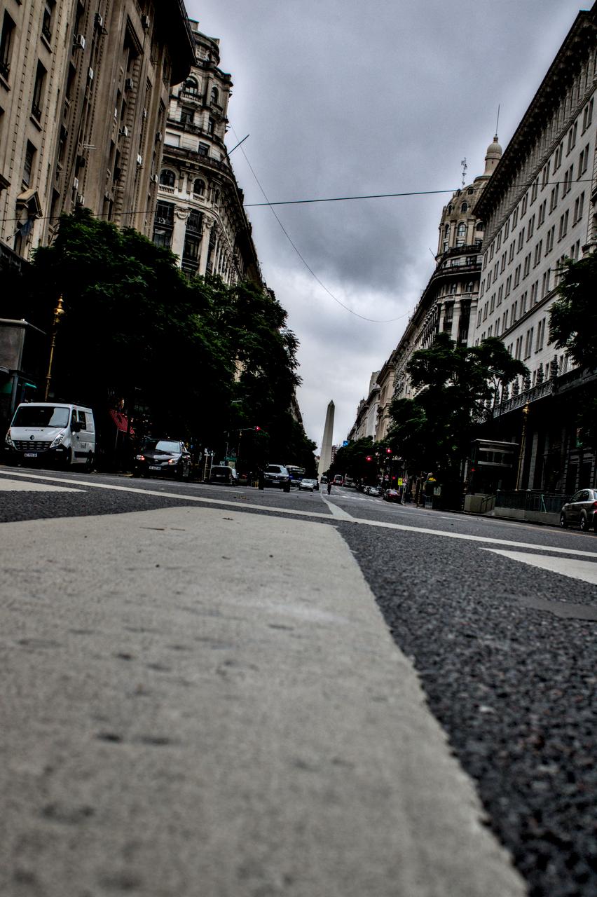 Foto inclinada; rua repleta de edifícios e obelisco ao fundo.