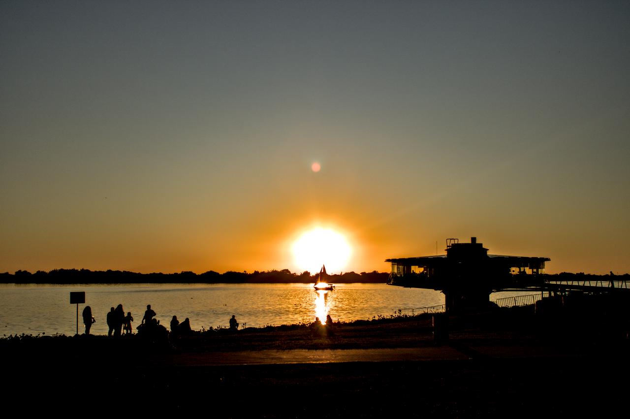 Sol poente atravessa veleiro diante de lago.