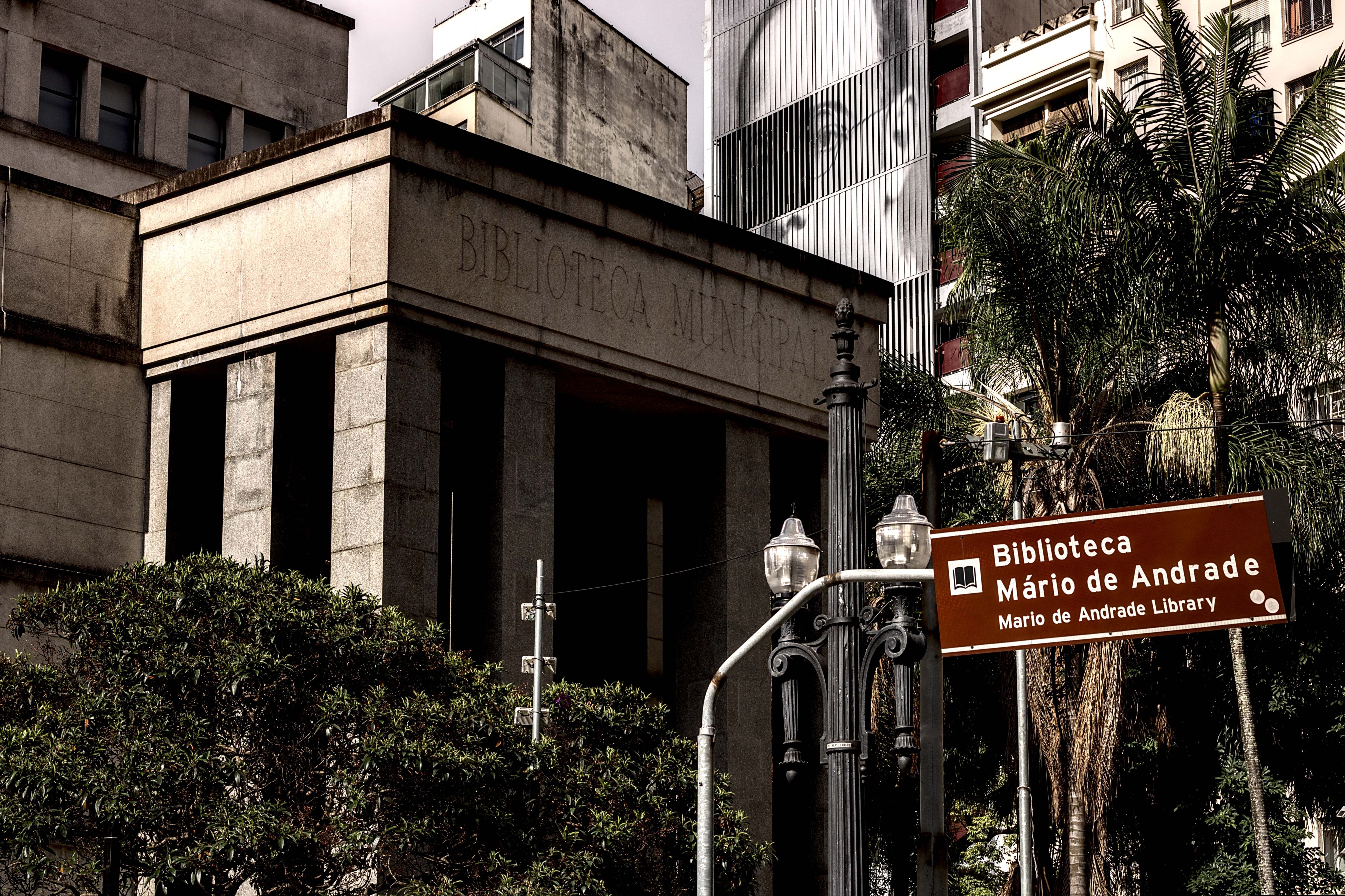 Pórtico cinzento de concreto. Ao fundo, retrato de Mário de Andrade em edifício. Num plano adiante, poste de luz da antiga Light Co.
