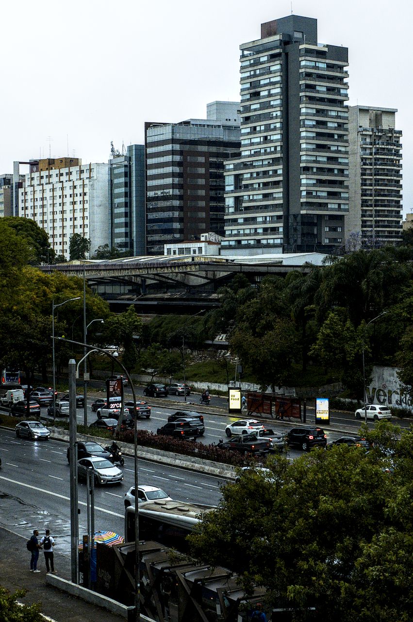 Vista para edifício horizontal entre avenida movimentada, em primeiro plano, e edifícios verticais, ao fundo.