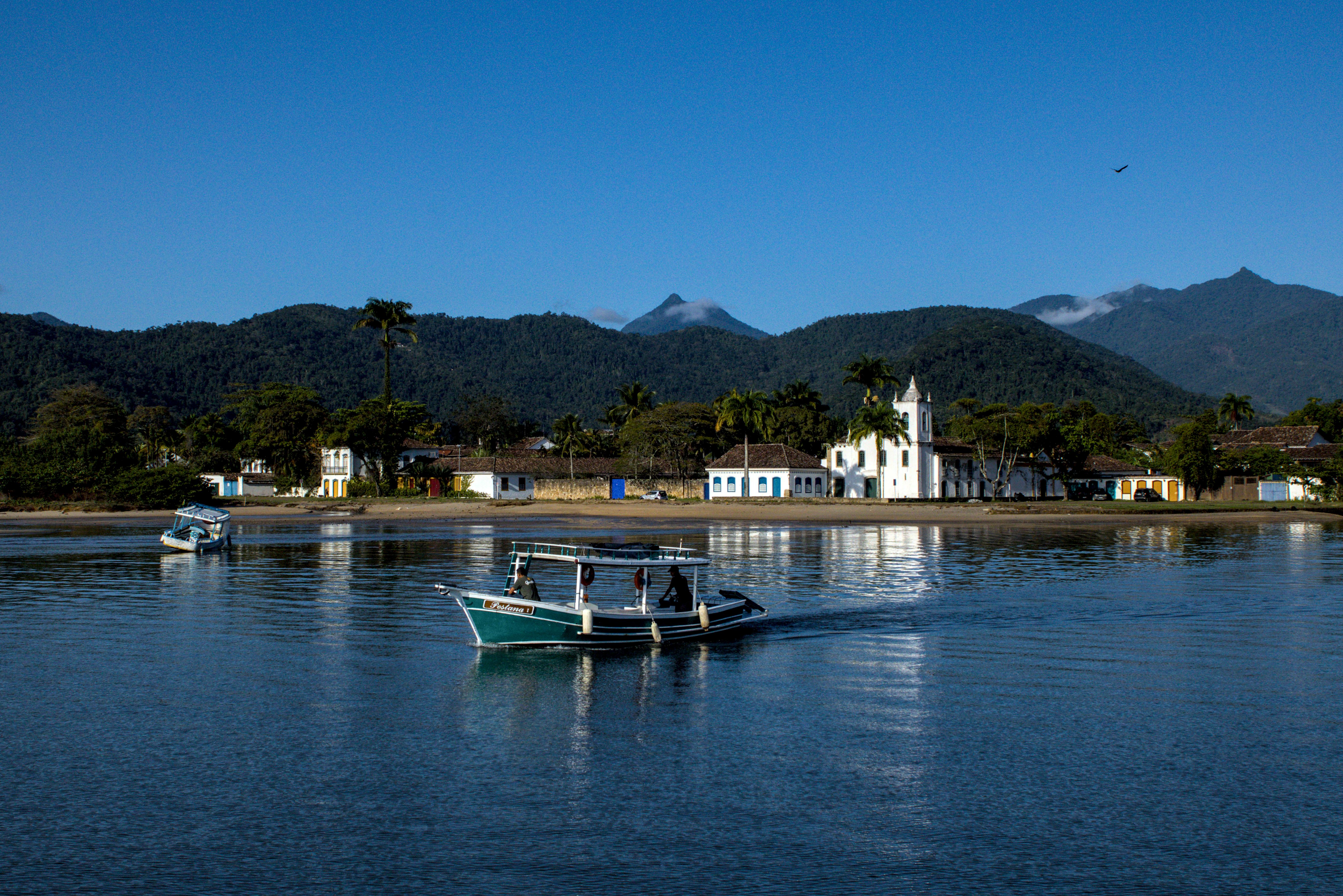 Serras de Paraty. Barco cruza rio em primeiro plano. Ao fundo, igreja branca e serras. Ave corta o céu azul.