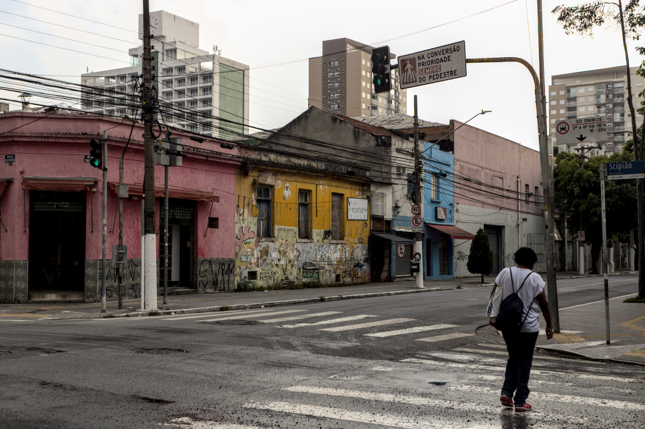 Pessoa atravessa a faixa de pedestres na rua molhada. Casario antigo e colorido na esquina oposta.