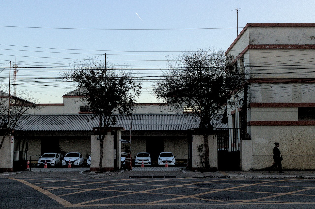 Costrução amarela clara e carros estacionados no interior do terreno. Dois terços de céu matinal com rastro de aeronave.