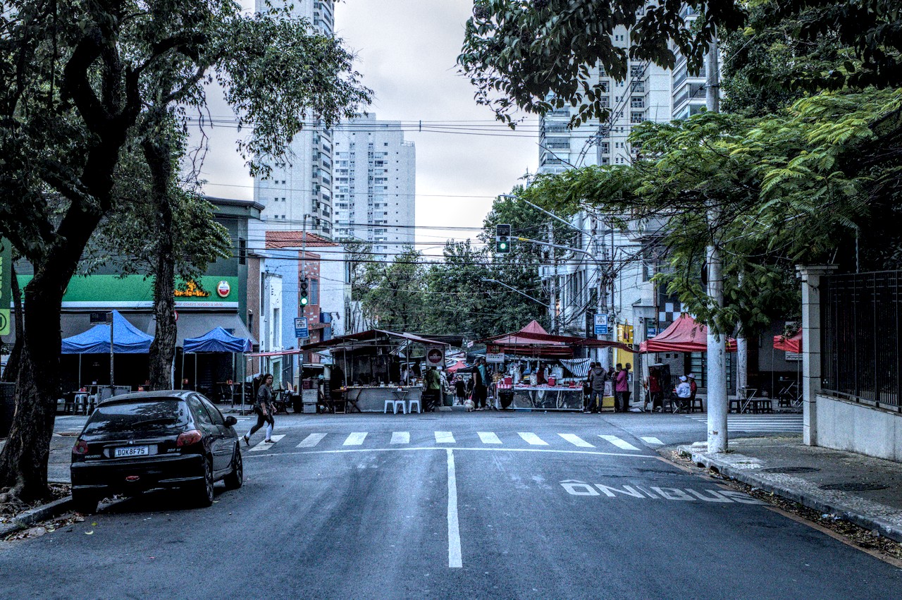 Rua com ladeira em direção a feira. Prédios ao fundo e árvores ao redor.