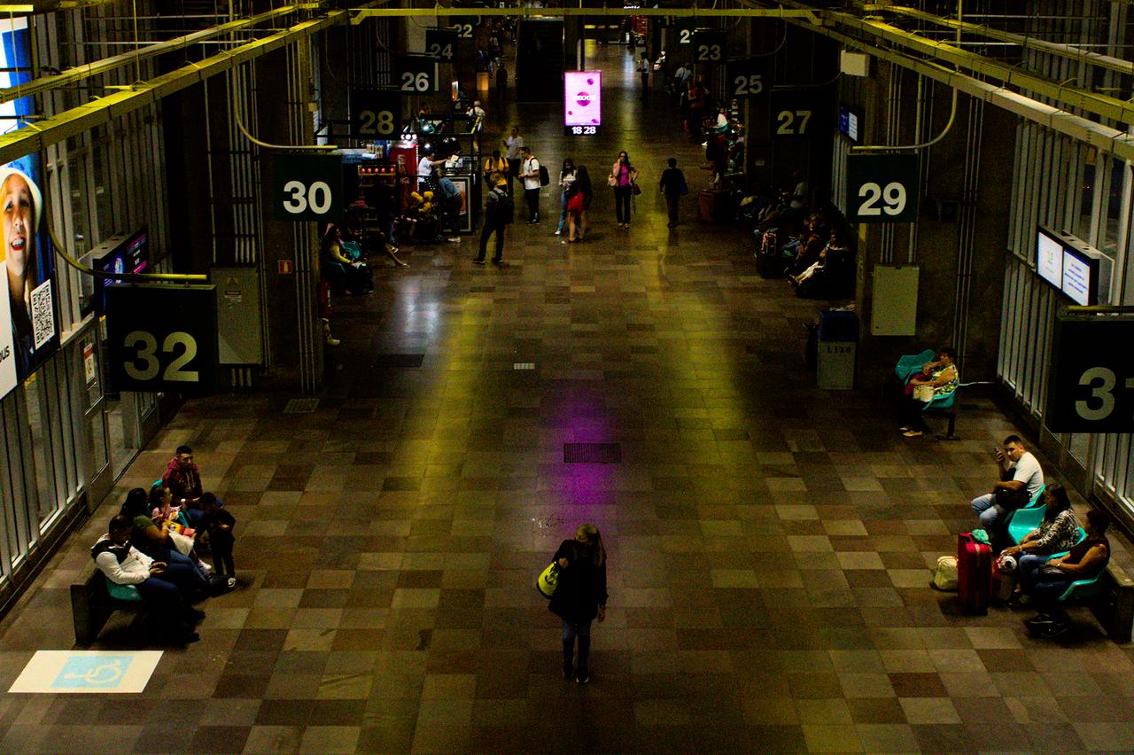 Vista de movimento de passageiros em rodoviária. Mulher caminha ao centro sob iluminação cor púrpura