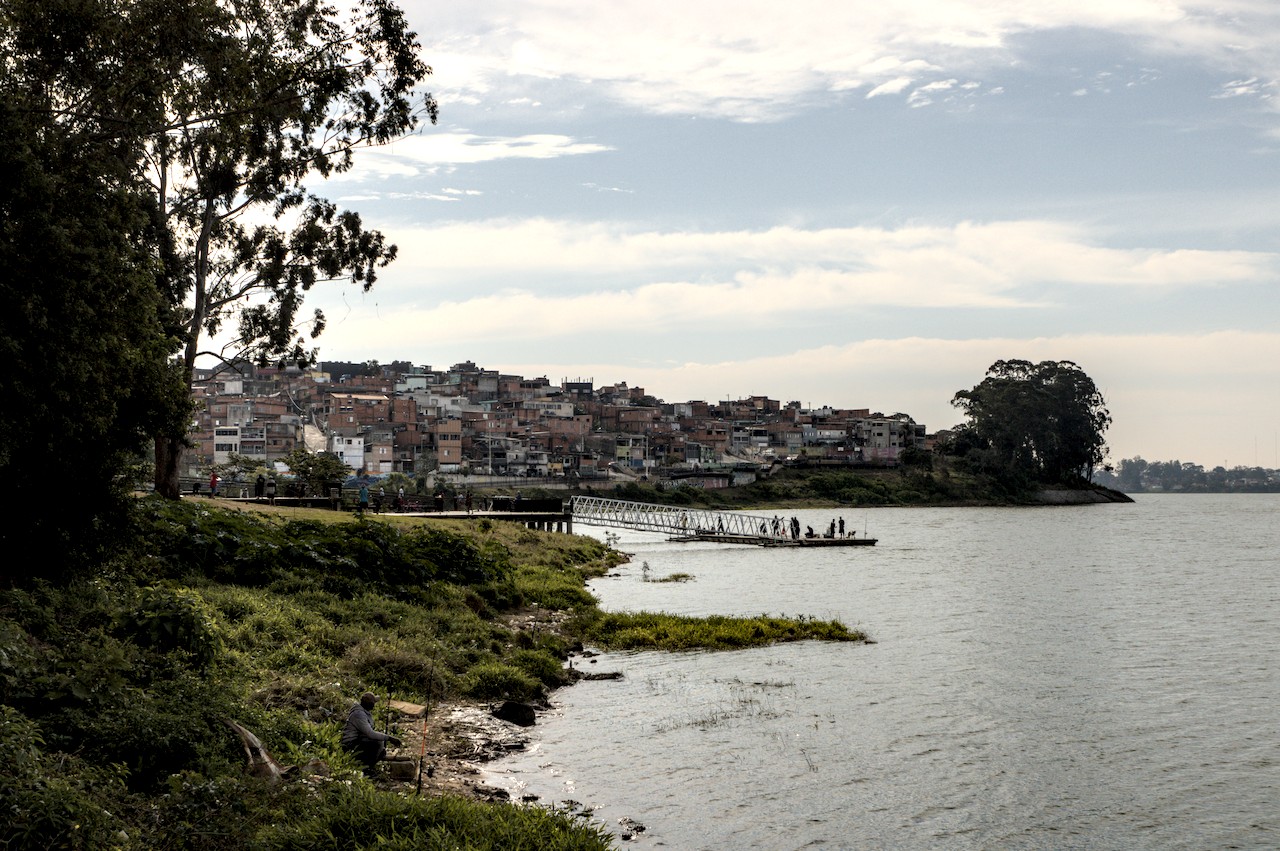 Vista ampla de parque com deque; em primeiro plano, idoso pesca sentado com duas varas; em segundo plano, deque de madeira com passarela onde famílias interagem; ao fundo, casario humilde.