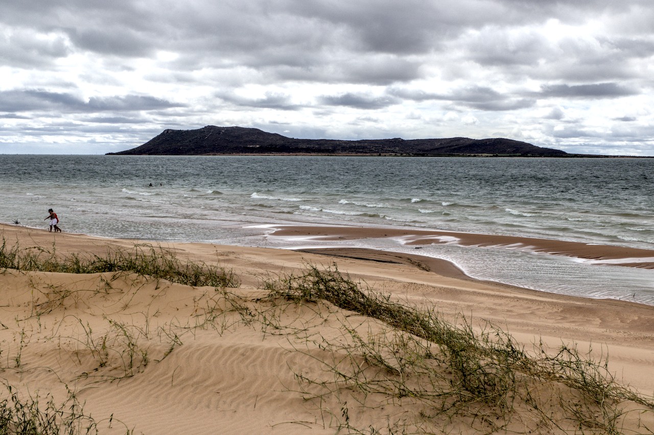 &ldquo;Praia de ilha em represa onde caminham duas pessoas. Porção de terra ao fundo.&rdquo;