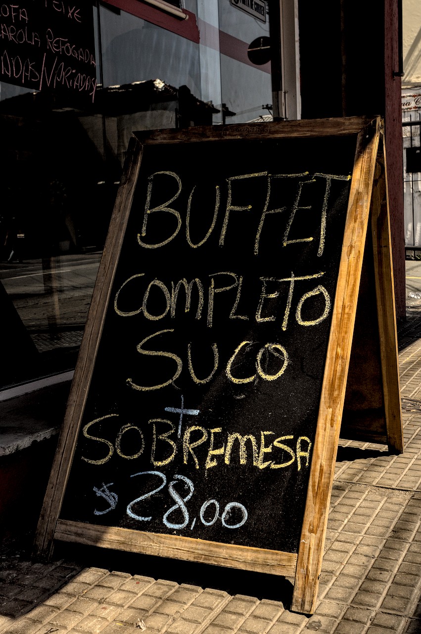 Lousa de cardápio. Lousa sob Sol duro com a frase 'Buffet Completo Suco + Sobremesa $28,00'.