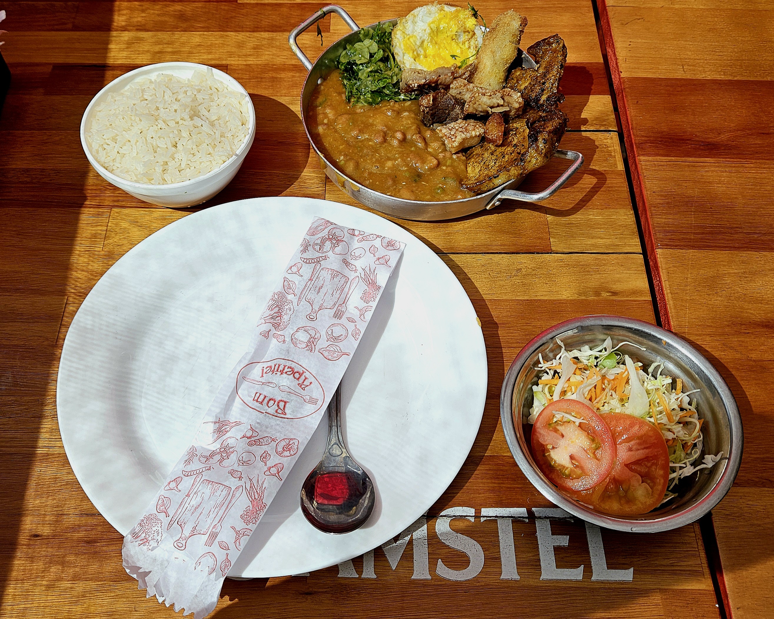 Prato com talheres, cuia de arroz, tijela de salada e panela com virado de feijão, bisteca com ovo, couve, banana empanada e torresmos.