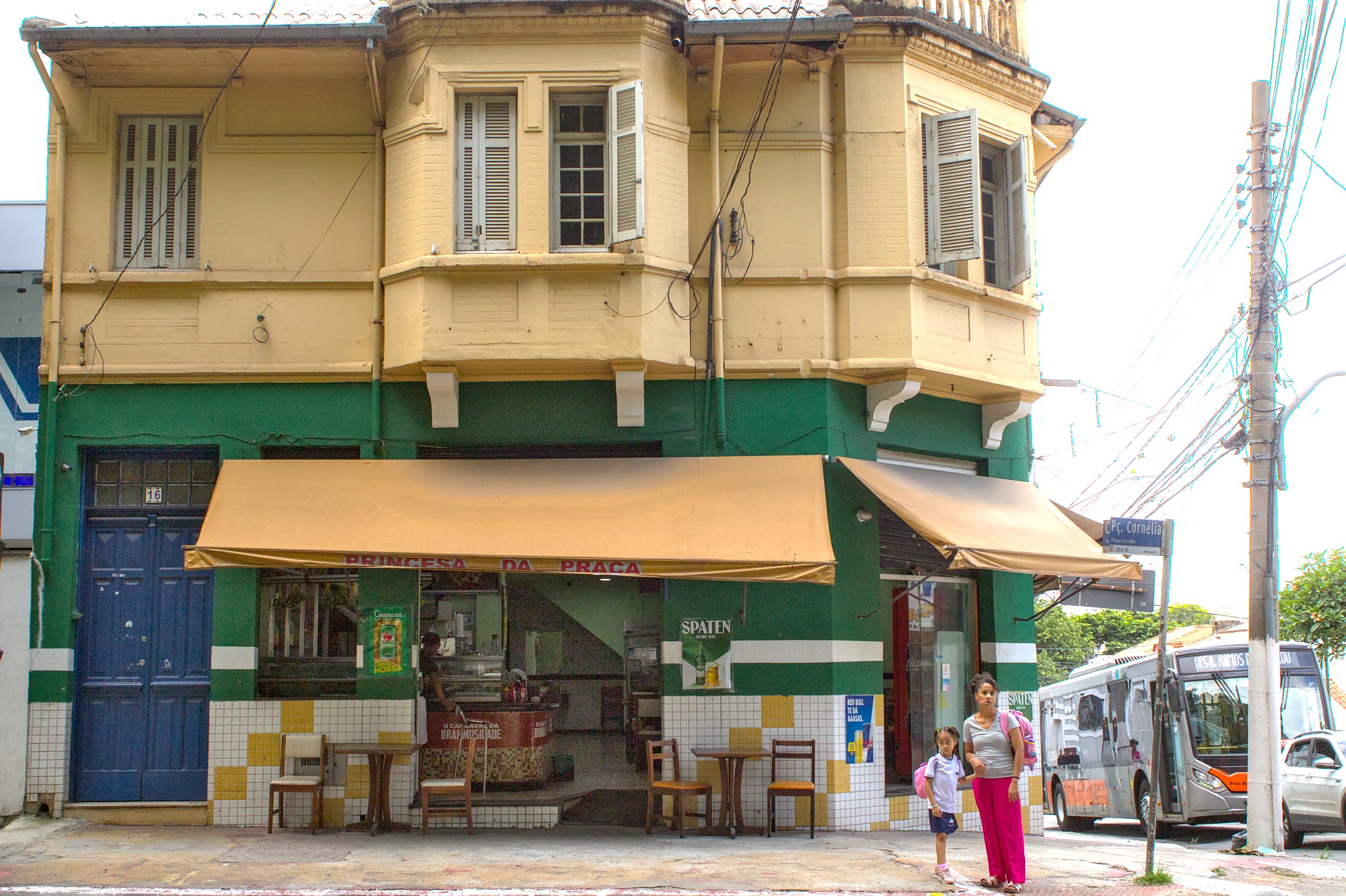 Boteco de esquina de fachada amarela. Pessoas aguardam para atravessar a rua.