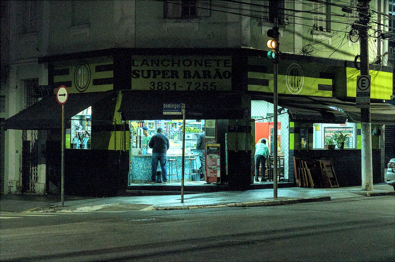 Boteco de esquina no escuro da madrugada; homem consome ao balcão.
