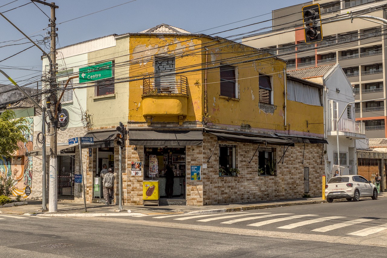 Boteco de esquina sob Sol duro do meio-dia; térreo em azulejos de cor pastel e primeiro andar degradado em cor amarela.