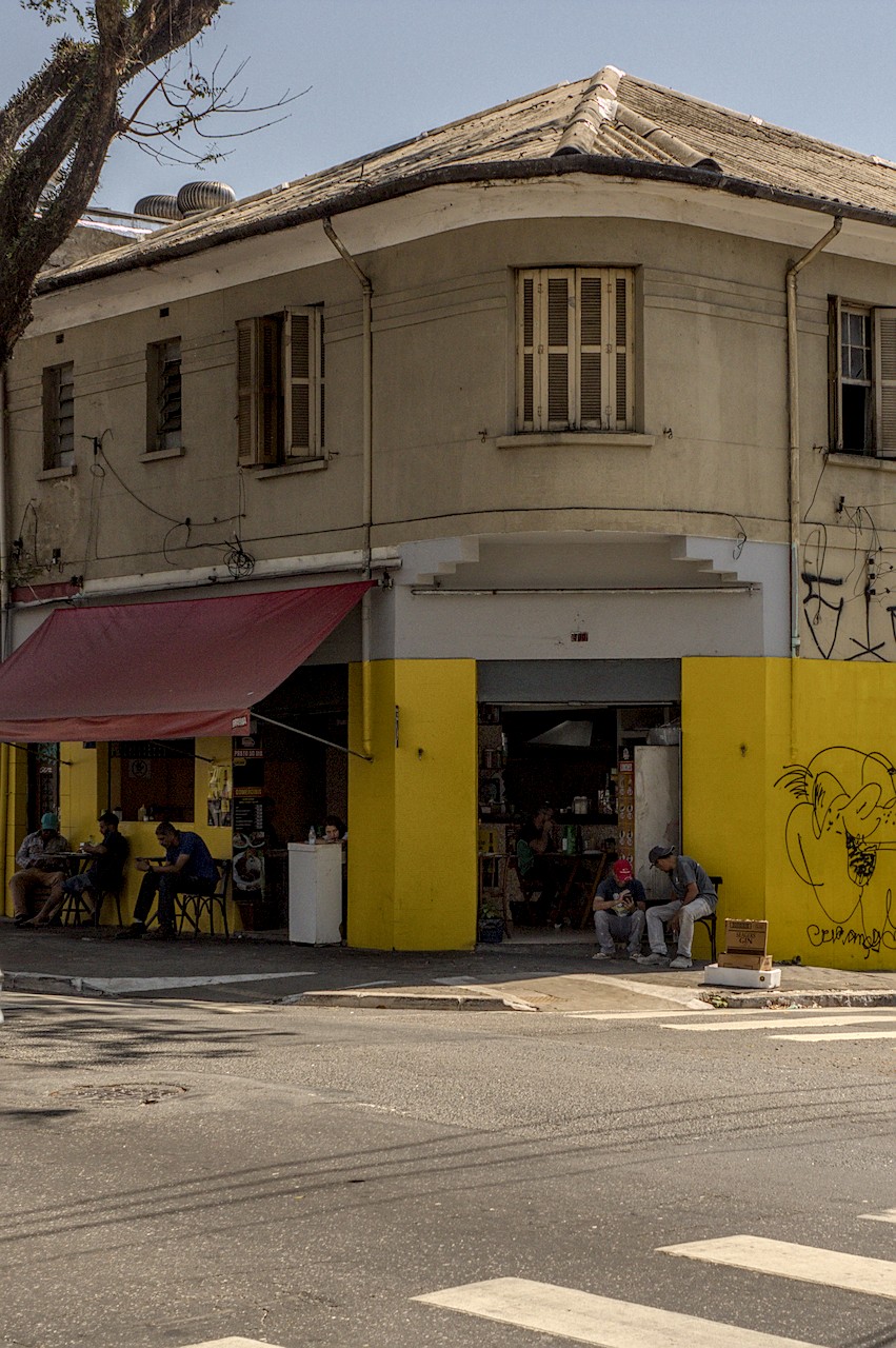 Boteco de esquina sob Sol duro do meio-dia; térreo amarelo, primeiro andar pastel; homens consomem na calçada.