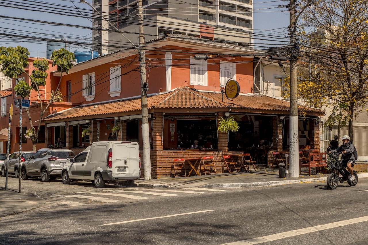 Boteco de esquina sob Sol duro do meio-dia; fachada cor-de-rosa e de tijolinhos; moto desco a rua à direita.