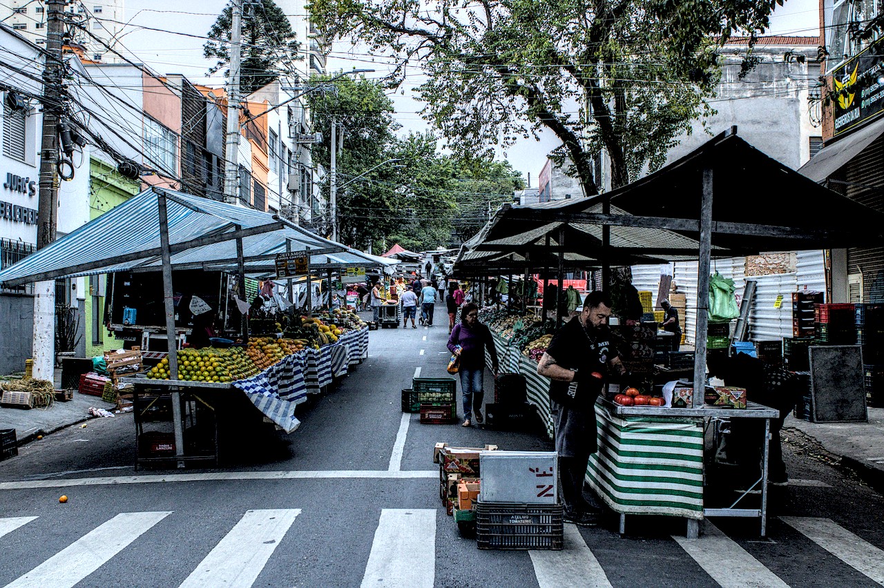 Barracas de hortifrútis em feira livre. Pessoas circulam.