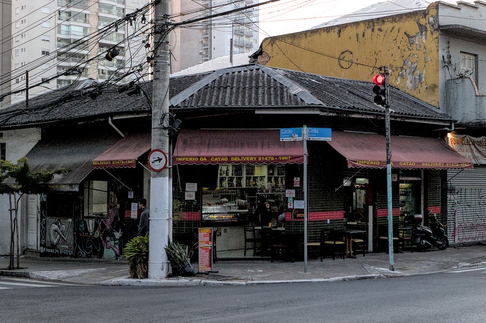 Boteco de esquina com mesas na calçada e moto estacionada.